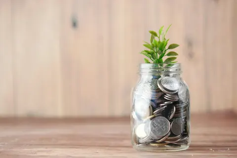 A glass jar filling with coins, a small plant growing beside it